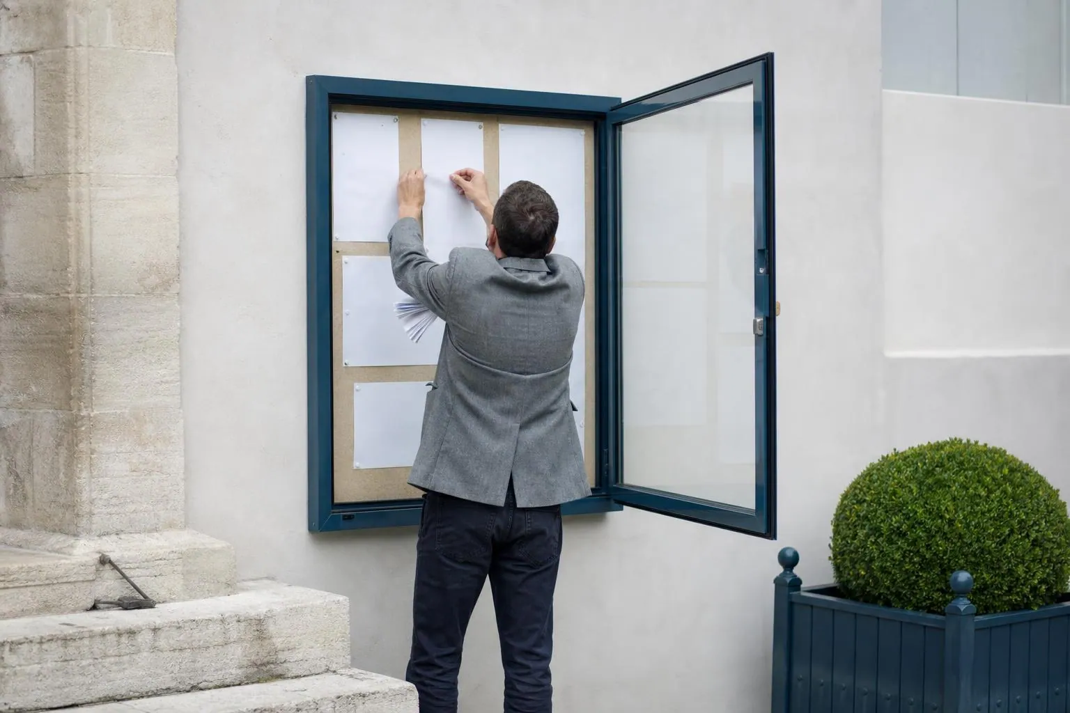Un agent municipal punaise des feuillets dans le panneau d’affichage vitré fixé sur la façade d’une mairie de village.