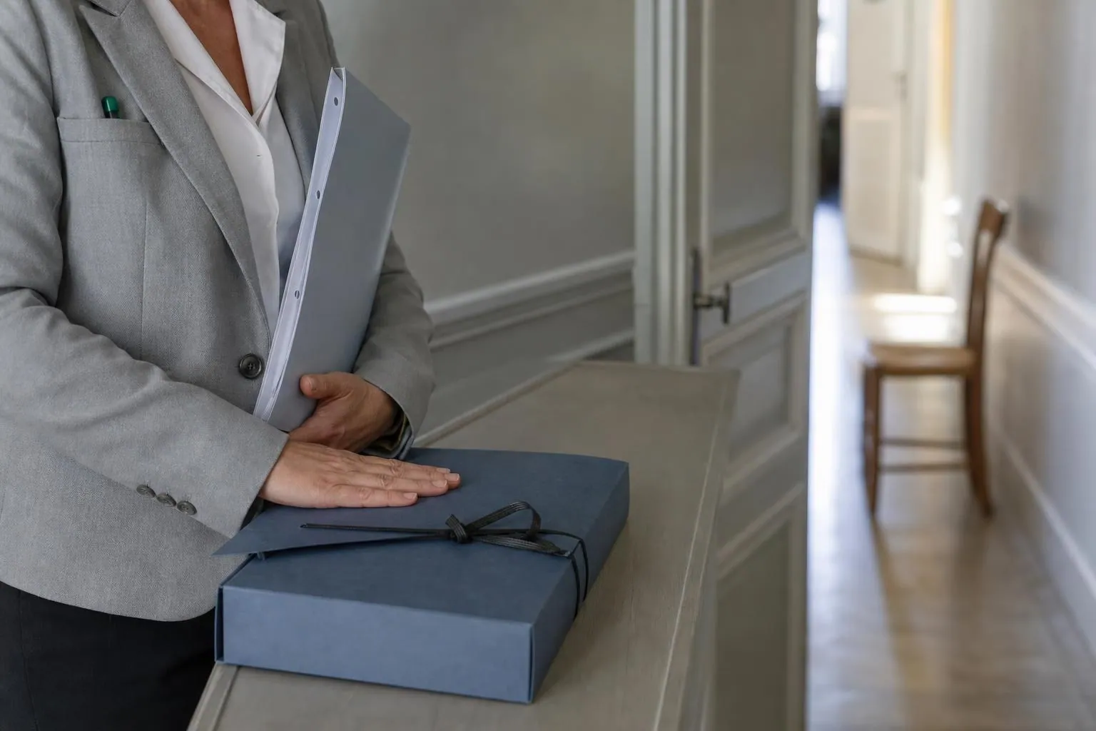 Une agente pose la main sur un parapheur fermé posé sur une console, devant la porte entrouverte du bureau du maire.