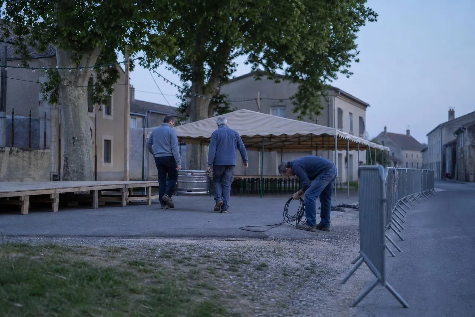Des bénévoles installent la buvette et les barrières d’une fête votive sur la place d’un village, à la tombée du jour.